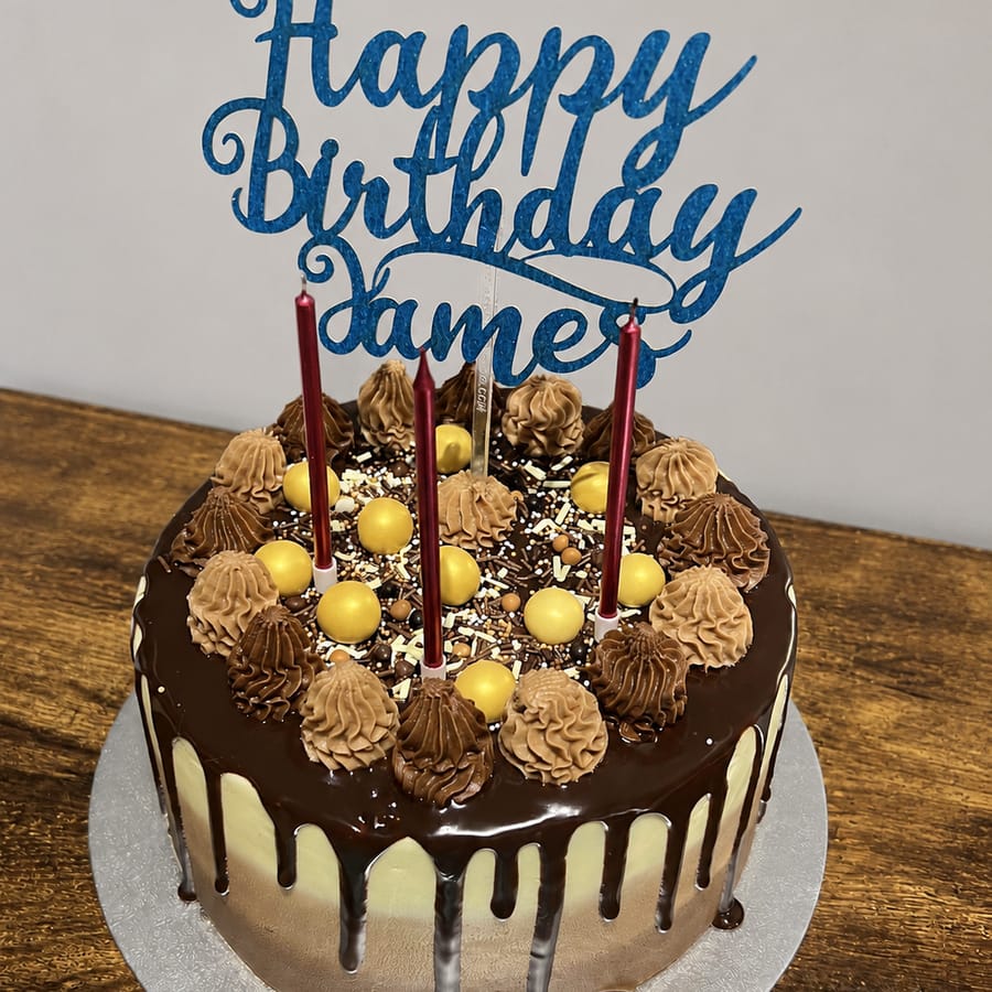 A loaded chocolate drip cake with chocolate swirls, gold pearls and a blue ‘Happy Birthday James’ topper