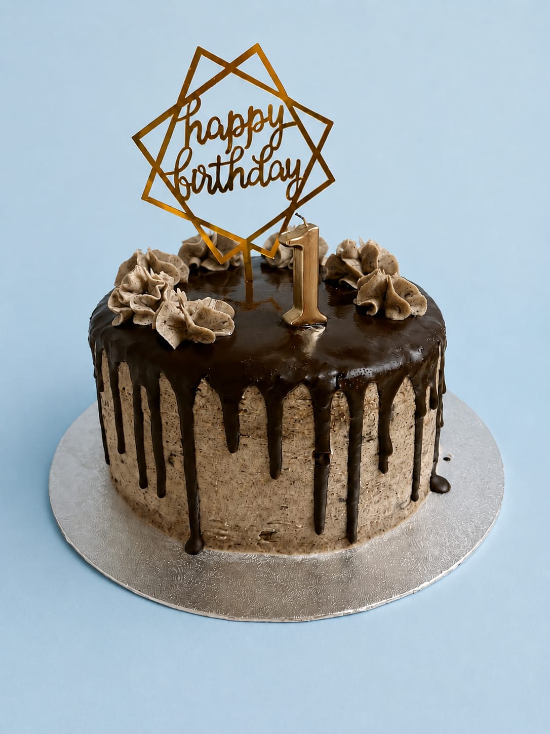 A chocolate drip celebration cake with a gold ‘happy birthday’ topper and a number 1 candle